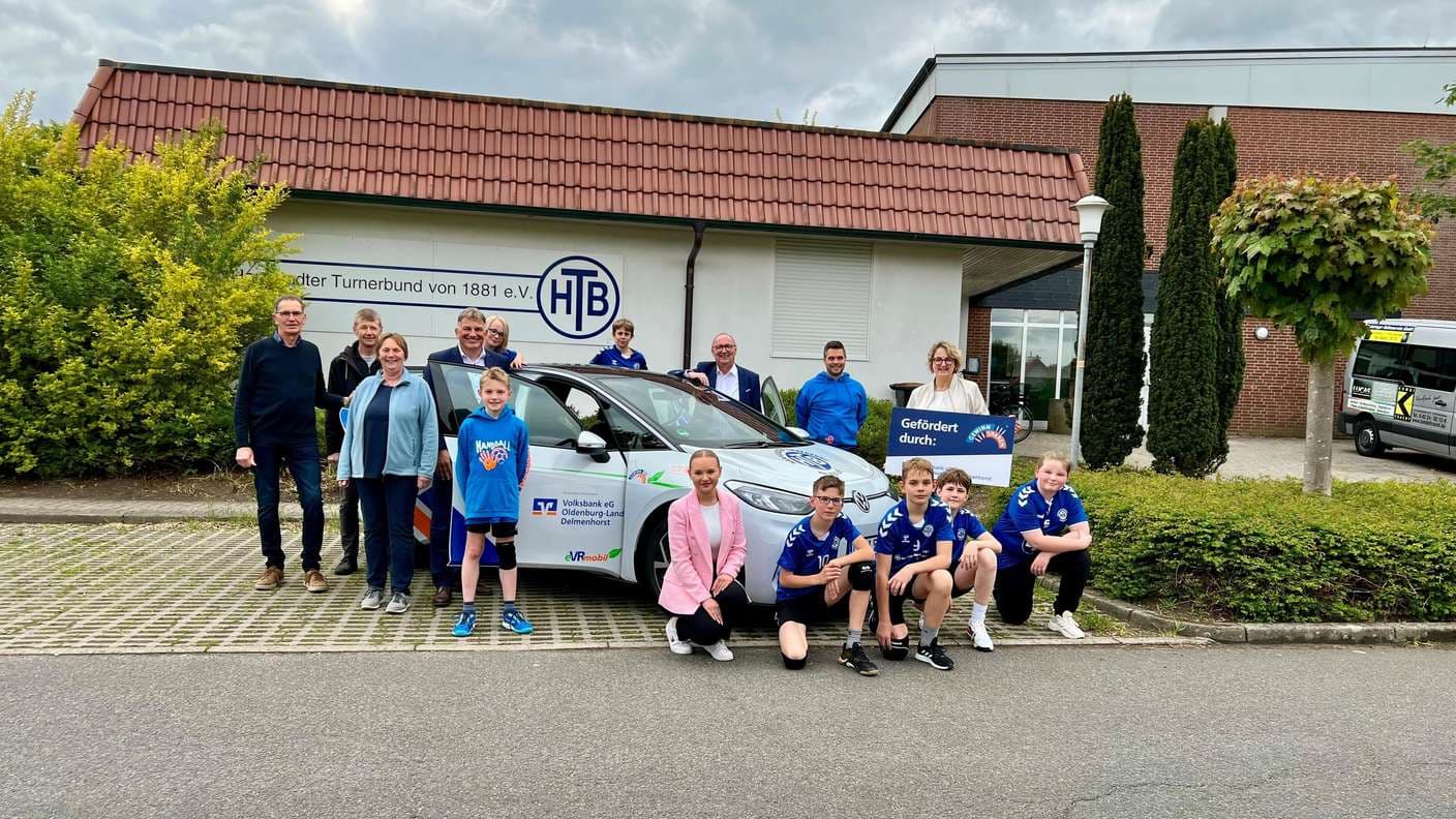 Gruppenfoto vor der HTB-Geschäftsstelle: Erwachsene und Kinder in blauen Handballtrikots um einen weißen VW ID.3 mit Volksbank-Aufschrift, eine Frau hält ein Schild „Gefördert durch“.